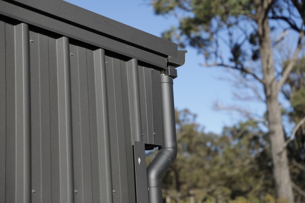 Close up of the corner of a dark grey steel shed with a gutter and downpipe system against a blue sky with eucalyptus trees out of focus in the background