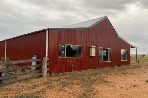 A red Australian style barn with grey trim and roof in a dusty rural setting with an overcast sky