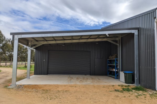 A front view of the lean to and open carport area to the left side of a larger grey shed which is just visible to the right
