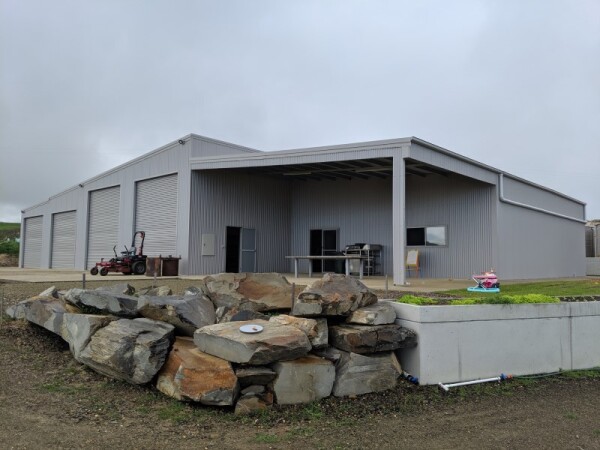 Tailor-made steel shed with four garage doors to the left,  an open bay on a lean-to to the right and a retaining wall and rock feature in the foreground