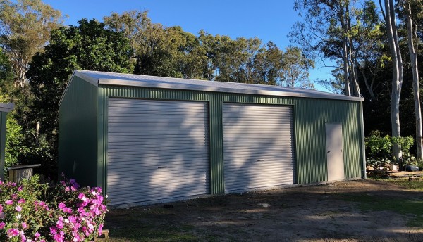 A green garage with white doors in a garden setting with purple flowers in the front left